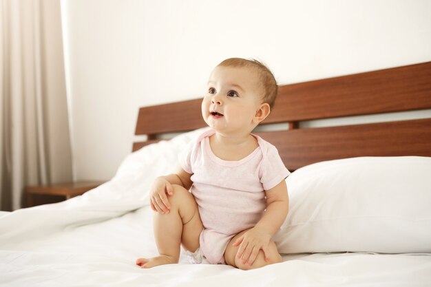 Retrato do bebê recém-nascido agradável bonito bonito na camisa cor-de-rosa que senta-se na cama em casa.