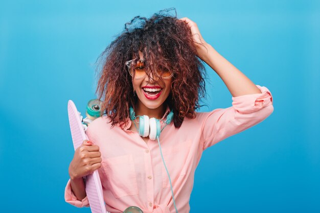 Retrato do close-up da espetacular mulata brinca com o cabelo castanho escuro com longboard. Mulher desportiva em fones de ouvido e camisa da moda de algodão, sorrindo na frente da parede azul.