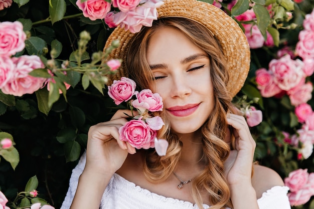 Foto grátis retrato do close-up da garota encaracolada em êxtase posando com rosas. tiro ao ar livre de uma mulher atraente com chapéu de palha, aproveitando o dia de verão.