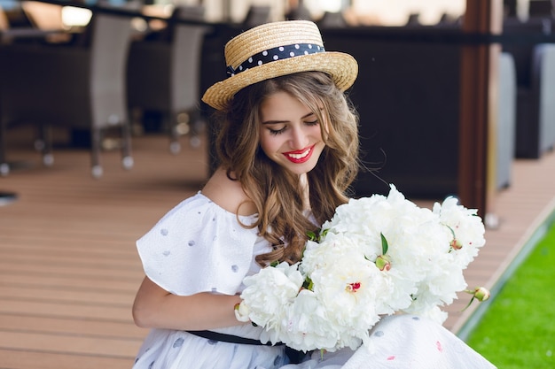 Retrato do close-up da linda garota com cabelo comprido no chapéu, sentado no chão, no terraço. ela usa um vestido branco com ombros nus, batom vermelho. ela tem flores brancas nas mãos.