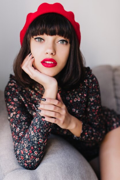 Retrato do close-up da magnífica menina morena com roupa francesa com maquiagem brilhante, posando no sofá. Mulher jovem elegante com cabelo curto escuro olha pensativamente para a câmera, apoiando o rosto com a mão.