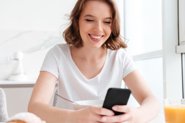 Retrato do close-up da mensagem de SMS alegre mulher no smartphone enquanto está sentado e tomando café na cozinha