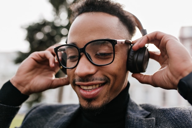 Retrato do close-up de adorável modelo masculino em óculos casuais relaxando com música no parque. Foto de um cara africano bonito com cabelo escuro posando em fones de ouvido