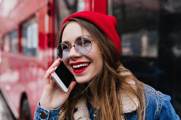 Retrato do close-up de incrível jovem com chapéu vermelho, falando no telefone na rua.