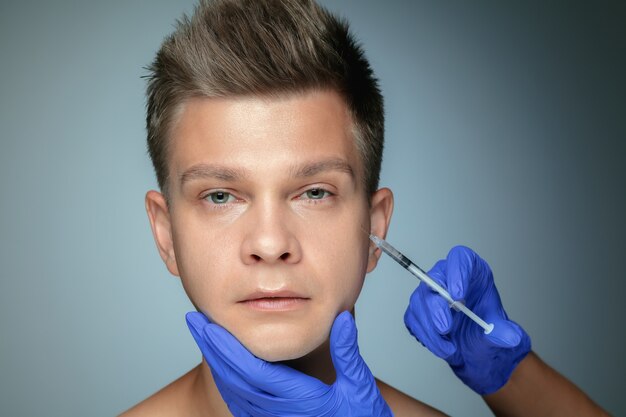 Retrato do close-up de jovem isolado na parede cinza. Procedimento de cirurgia de enchimento. Conceito de saúde e beleza masculina, cosmetologia, autocuidado, cuidados com o corpo e a pele. Anti-envelhecimento.