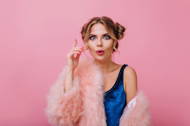 Retrato do close-up de menina encaracolada surpresa em roupas de veludo engraçadas posando com a mão para cima. Mulher jovem loira incrível com maquiagem na moda se lembrou de algo importante, isolado no fundo rosa.