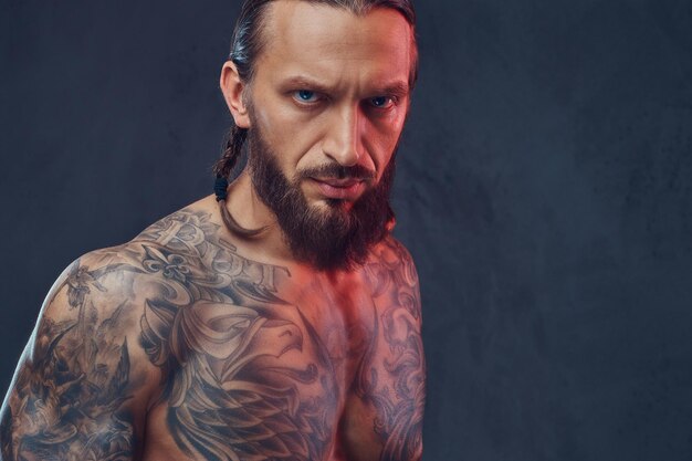 Retrato do close-up de um homem tatuado barbudo musculoso com um corte de cabelo elegante, isolado em um fundo escuro.