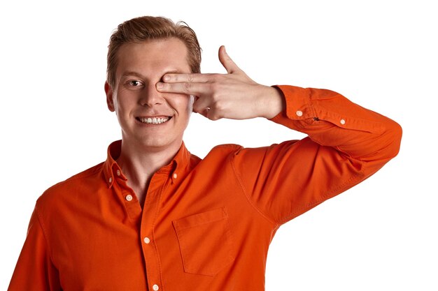 Retrato do close-up de um jovem adorável gengibre em uma elegante camisa laranja, fechando o olho com os dedos enquanto posava isolado no fundo branco do estúdio. Expressões faciais humanas. Conceito de emoções sinceras