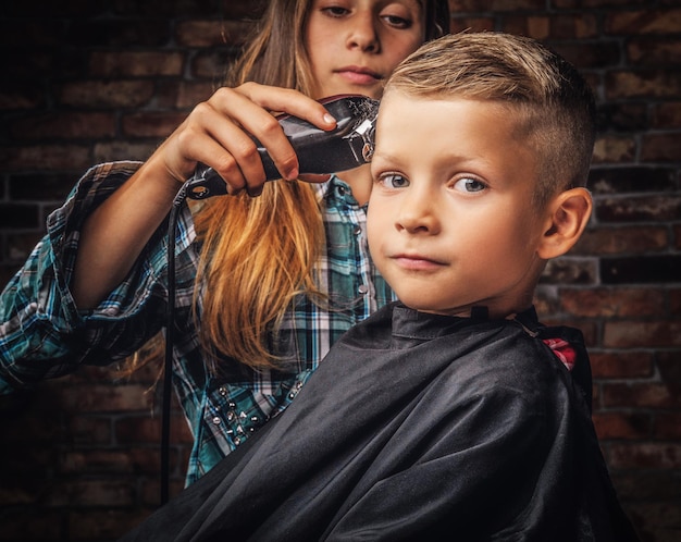 Retrato do close-up de um menino bonito da pré-escola cortando o cabelo. A irmã mais velha corta seu irmãozinho com um aparador contra uma parede de tijolos.