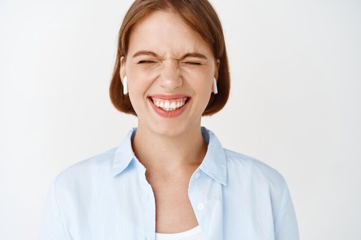 Foto grátis retrato do close-up de uma garota animada, sorrindo com os olhos fechados, curtindo a música em fones de ouvido sem fio, em pé na parede branca