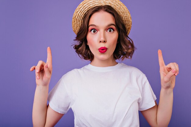 Retrato do close-up de uma garota de olhos escuros surpresa com chapéu de verão. Foto interna do modelo feminino encaracolado engraçado em t-shirt branca, posando com os dedos na parede roxa.