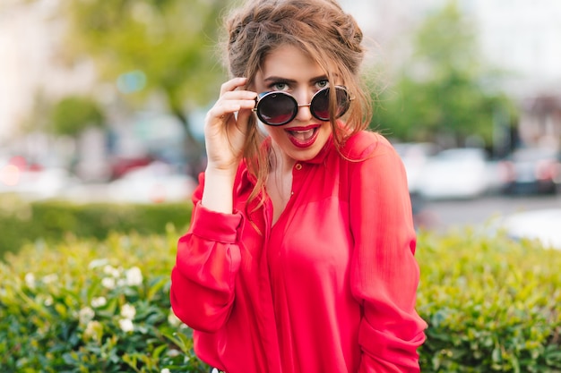 Retrato do close-up de uma linda garota de óculos escuros, posando para a câmera no parque. Ela usa blusa vermelha e um belo penteado. Ela está olhando para a câmera.