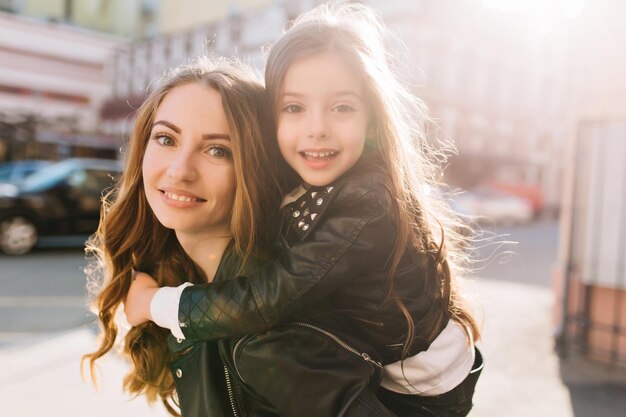 Retrato do close-up de uma linda mulher bonita e sua filha encantadora com grandes olhos escuros, posando no meio urbano. Menina linda morena montada nas costas da mãe e abraçando-a suavemente com um sorriso