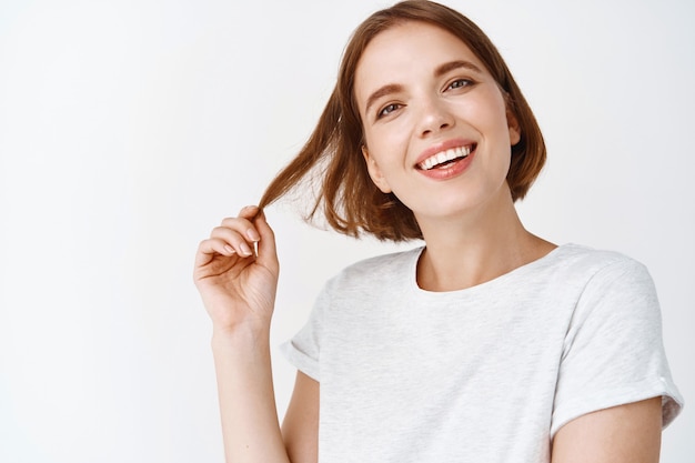 Retrato do close-up de uma menina bonita com cabelo curto, sorrindo com os dentes brancos e parecendo feliz, brincando com o corte de cabelo, em pé contra uma parede branca