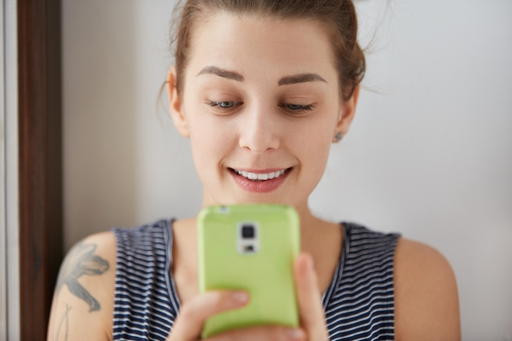 Foto grátis retrato do close-up de uma menina europeia jogando em seu smartphone verde. mulher jovem e encantadora segurando seu gadget com as duas mãos, olhando para o visor com um leve sorriso. tiro de luz do dia dentro de casa.