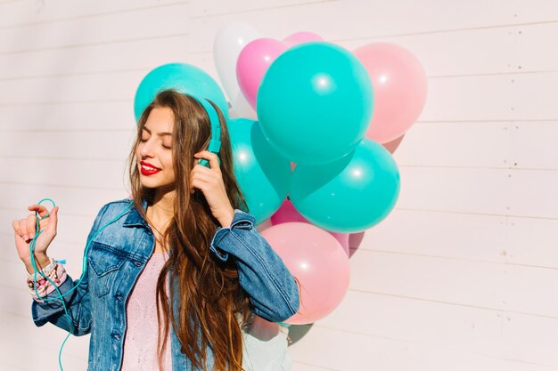 Retrato do close-up de uma menina morena de cabelos compridos no elegante casaco jeans, para a festa de aniversário. Mulher jovem e atraente posando com balões brilhantes enquanto ouve música em fones de ouvido com os olhos fechados