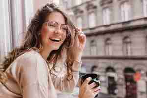 Foto grátis retrato do close-up de uma menina morena sorridente com um suéter bege tomando café no fundo da cidade