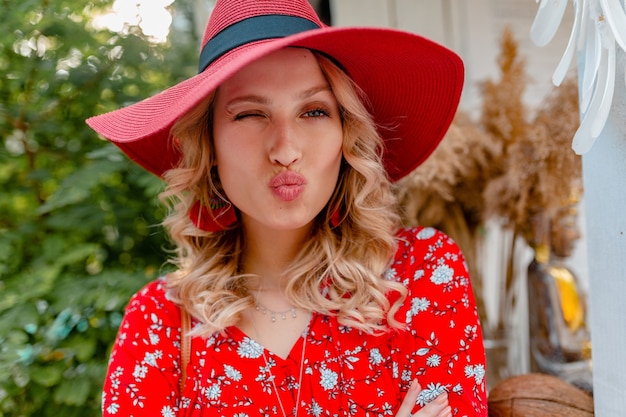 Retrato do close-up de uma mulher loira elegante e atraente sorridente com chapéu vermelho palha e blusa, roupa de verão, cara engraçada beijo