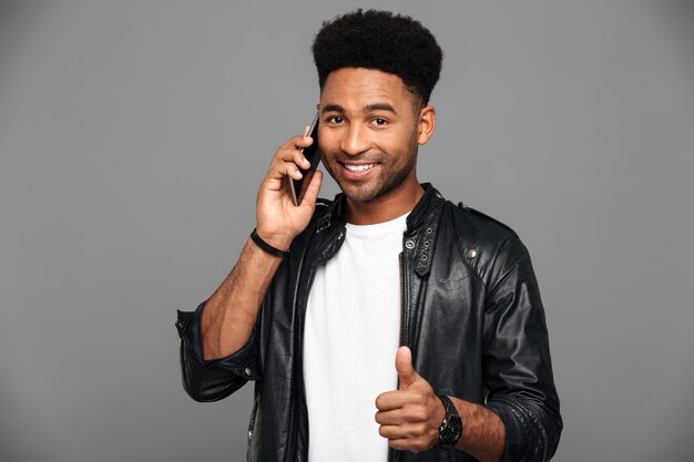 Retrato do close-up do homem afro-americano elegante sorridente, falando no celular enquanto mostra o polegar para cima gesto, olhando