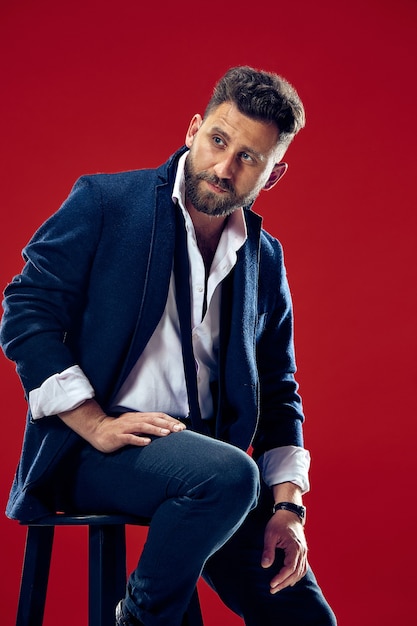 Retrato do conceito de beleza masculina de um jovem elegante com um corte de cabelo estiloso, vestindo um terno da moda posando sobre a parede vermelha