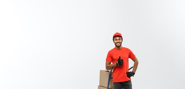 Retrato do conceito de entrega de um entregador afro-americano bonito ou mensageiro empurrando o caminhão de mão com pilha de caixas isoladas no espaço de cópia de fundo do estúdio cinza