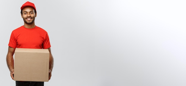 Foto grátis retrato do conceito de entrega de um entregador afro-americano feliz em pano vermelho segurando um pacote de caixa isolado no espaço de cópia de fundo do estúdio cinza