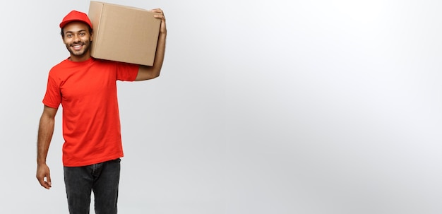 Foto grátis retrato do conceito de entrega de um entregador afro-americano feliz em pano vermelho segurando um pacote de caixa isolado no espaço de cópia de fundo do estúdio cinza