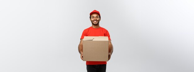 Retrato do conceito de entrega de um entregador afro-americano feliz em pano vermelho segurando um pacote de caixa isolado no espaço de cópia de fundo do estúdio cinza