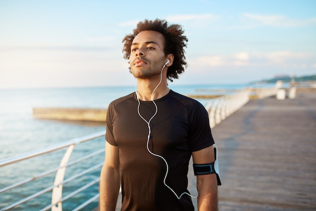 Foto grátis retrato do corredor homem musculoso de pele escura apto com penteado espesso olhando concentrado em roupas esportivas pretas, usando fones de ouvido brancos.