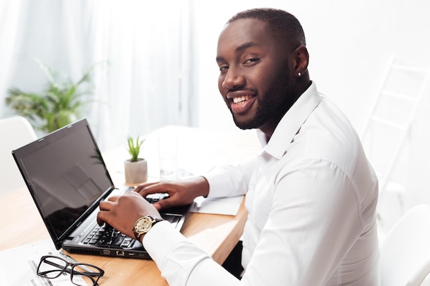 Retrato do empresário americano africano sorridente na camisa branca sentado e olhando alegremente na câmera enquanto trabalhava em seu laptop no escritório isolado