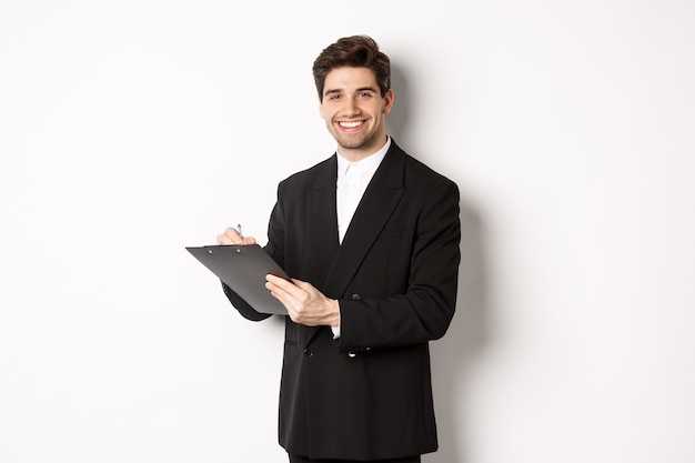 Retrato do empresário confiante em terno preto, assinando documentos e sorrindo, feliz em pé contra um fundo branco.