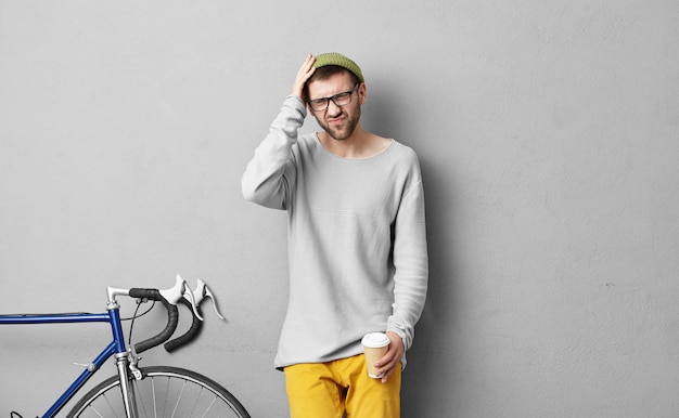 Retrato do estilo de vida de moda jovem macho com barba, tendo um olhar doloroso por causa de dor de cabeça, isolado na parede cinza com bicicleta fixa e segurando papercup, bebendo café para ir