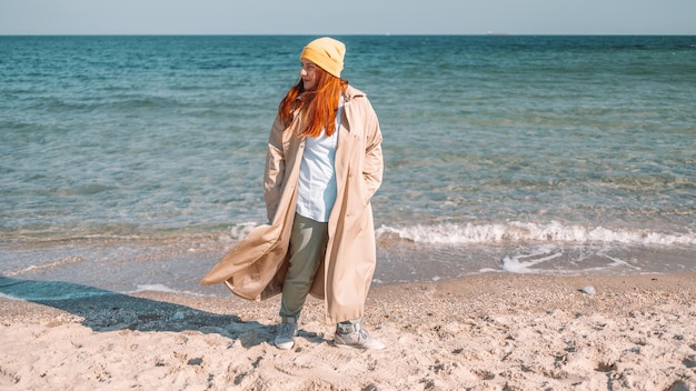 Retrato do estilo de vida de uma elegante mulher de cabelo ruivo caucasiana que caminha ao longo da praia perto do mar em um dia ensolarado