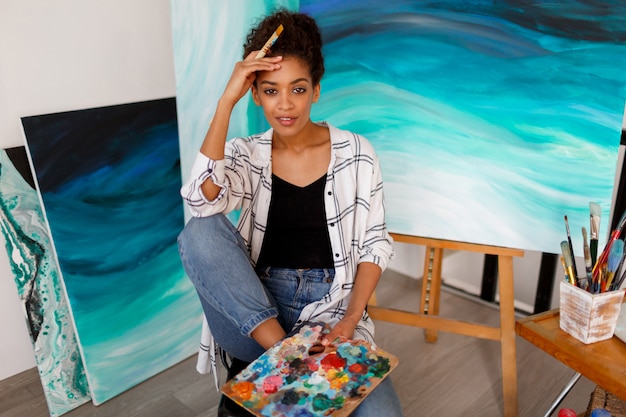 Retrato do estilo de vida do jovem estudante africano sentado com incrível arte abstrata mar acrílico mão desenhada obras de arte no estúdio.