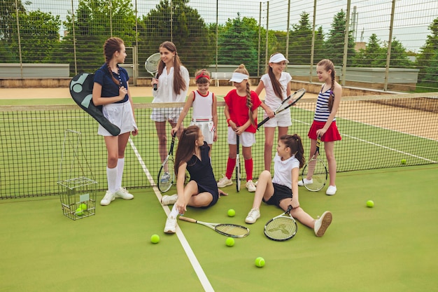 Retrato do grupo de meninas como jogadores de tênis que mantêm raquetes de tênis contra a grama verde da corte ao ar livre. Elegantes jovens adolescentes posando no parque. Estilo de esporte. Conceito de moda adolescente e crianças.