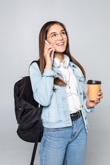 Foto grátis retrato do lado da mulher jovem estudante falar com smartphone, segurando o café para ir copo isolado na parede cinza