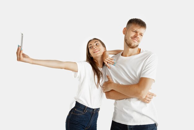 Retrato do lindo casal jovem isolado no fundo branco do estúdio. Expressão facial, emoções humanas, conceito de publicidade. Mulher fazendo selfie ou vídeo para vlog, homem fica entediado, não quer fazer.