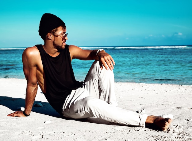 Foto grátis retrato do modelo de homem bonito moda banhos de sol hipster vestindo roupas casuais em camiseta preta e óculos de sol, sentado na areia branca, perto do céu e oceano azul