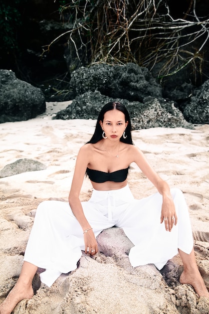 Retrato do modelo de mulher bonita caucasiano com cabelos longos escuros nas calças clássicas de pernas largas, sentado na praia de verão com areia branca perto de pedras