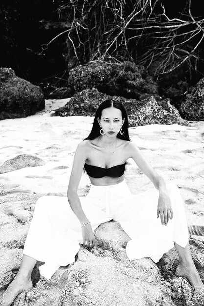 retrato do modelo de mulher bonita caucasiano com cabelos longos escuros nas calças clássicas de pernas largas, sentado na praia de verão com areia branca perto de pedras