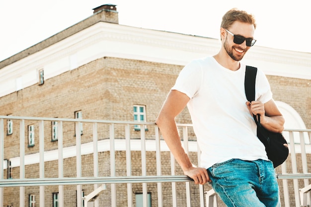 Retrato do modelo lambersexual de hipster sorridente bonito homem elegante vestido com camiseta branca e jeans moda masculina posando no fundo da rua perto da cerca na praia