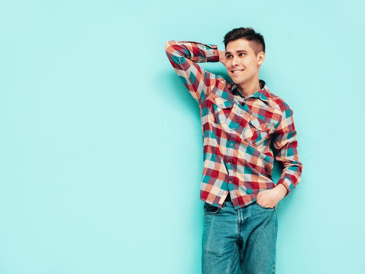 Foto grátis retrato do modelo sorridente bonito sexy homem elegante vestido com camisa xadrez vermelha e jeans moda hipster masculino posando perto de azul cinza no estúdio isolado