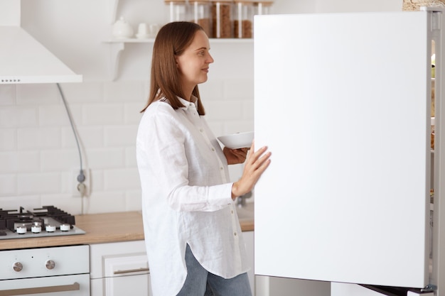 Retrato do perfil da bela jovem adulta vestindo camisa branca, olhando sorrindo dentro da geladeira com um sorriso agradável, segurando o prato nas mãos, posando com cozinha em segundo plano.