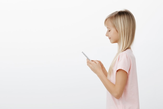 Retrato do perfil de alegre calma garota atraente com cabelo loiro em uma camiseta rosa, segurando um smartphone, sorrindo para a tela, enviando foto para a mãe por meio de mensagens, dizendo à mãe que ela terminou as tarefas domésticas