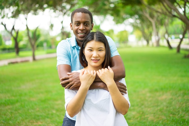 Retrato dos pares multi-étnicos novos felizes que abraçam e que sorriem no parque.