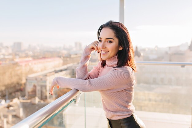 Retrato elegante sorriu jovem morena no terraço em um dia ensolarado na vista da cidade grande. Sorrindo para o lado, sucesso, tempo de relaxamento, relaxando na luz do sol.