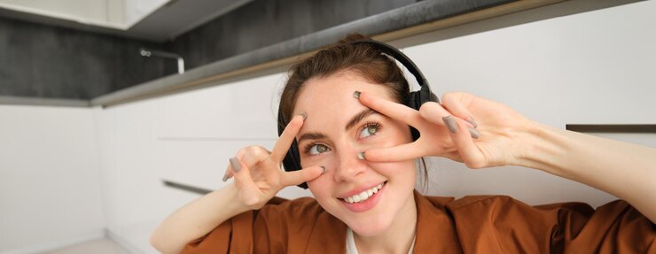 Foto grátis retrato em close-up de mulher com fones de ouvido mostra paz vsigns perto dos olhos sorri e parece feliz senta-se em