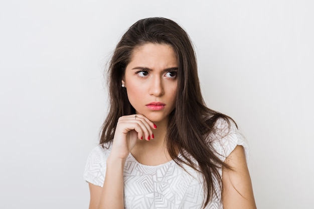 Retrato em close-up de uma jovem mulher pensando em uma blusa branca, carrancuda, segurando a mão em seu rosto, olhando para o lado, expressão facial séria, tendo um problema isolado