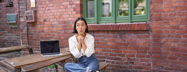 Retrato em tamanho real de uma menina asiática sentada com laptop parece espantado e surpreso e espia a tela