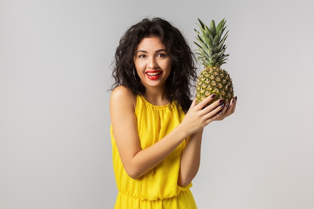 Retrato emocional de uma jovem mulher exótica morena de vestido amarelo, segurando abacaxi, expressão de carinha engraçada, emoção positiva, isolado, frutas tropicais, dieta, feliz, sorridente, estilo de vida saudável